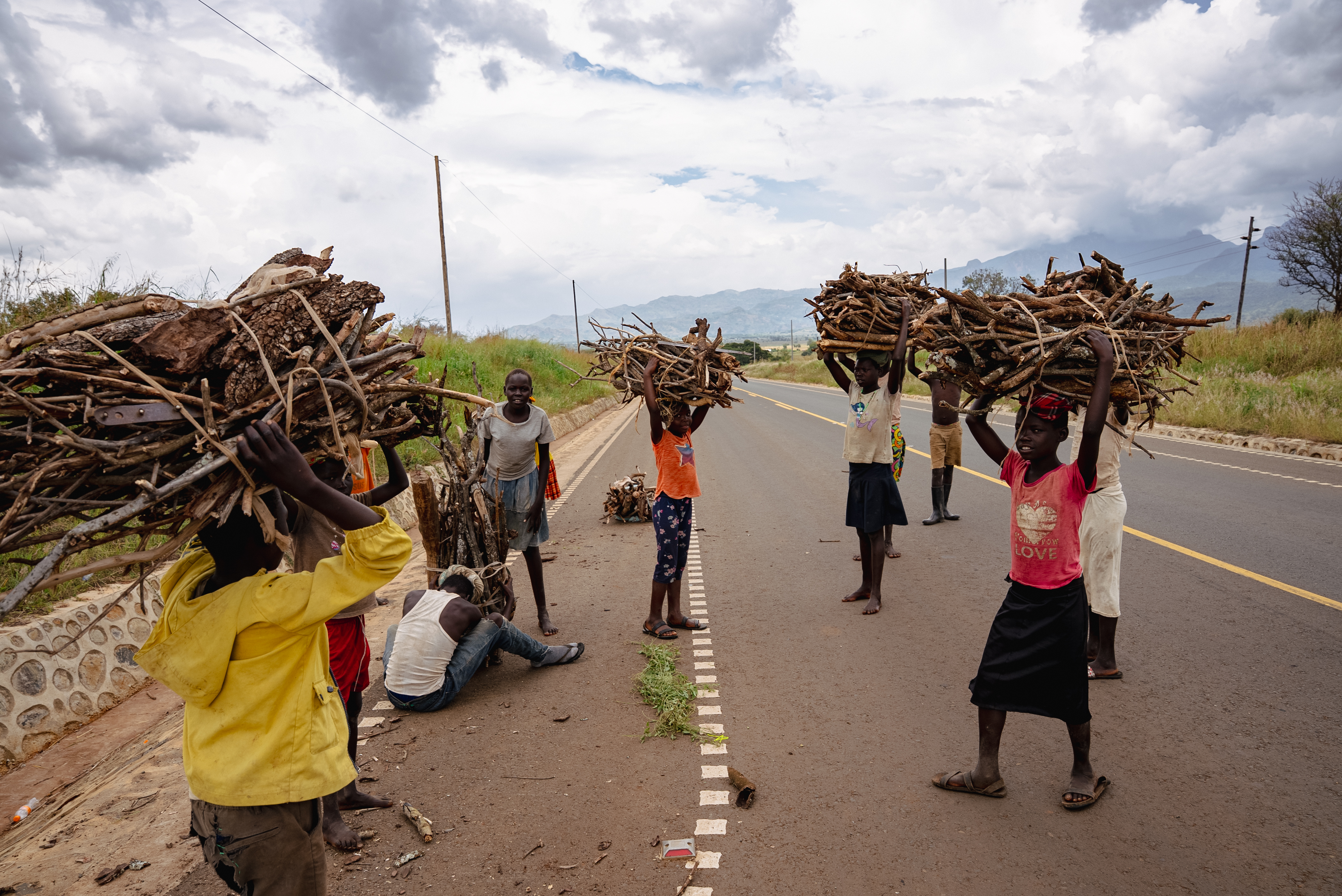 Children carrying bundles of firewood along the roadside beneath the mountains
